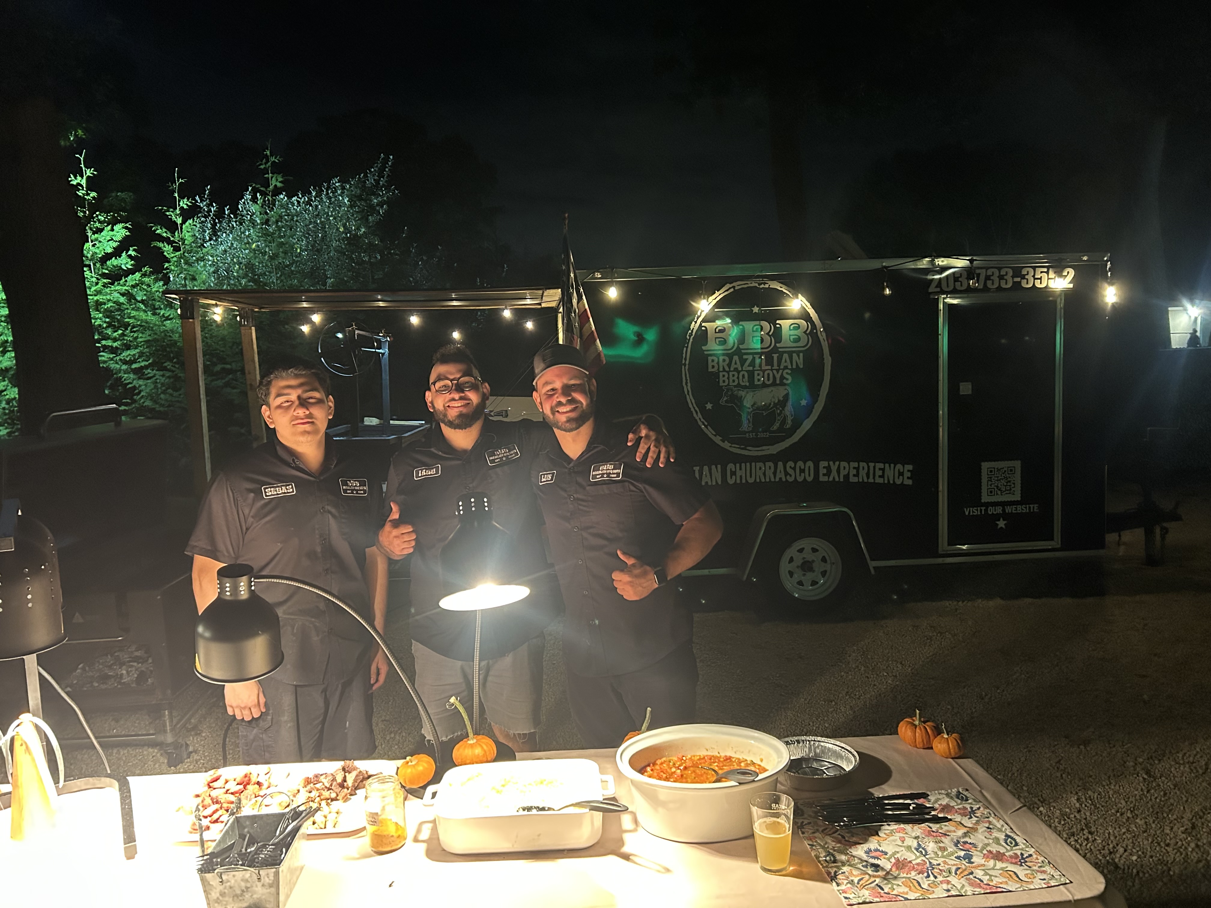 The Brazilian BBQ Boys team posing together in front of their catering trailer at a nighttime event