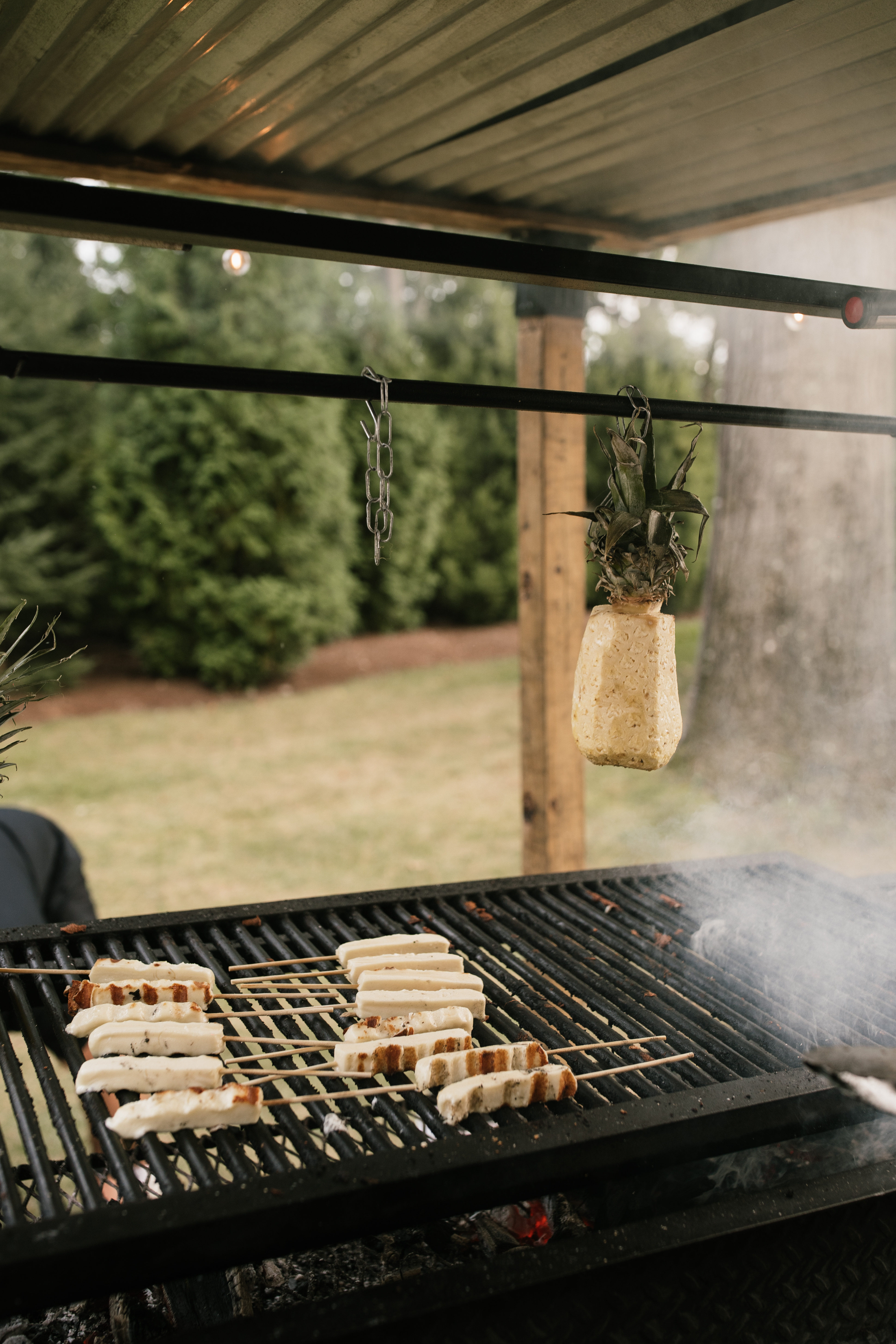 Queijo coalho cheese skewers grilling over charcoal with pineapple hanging above